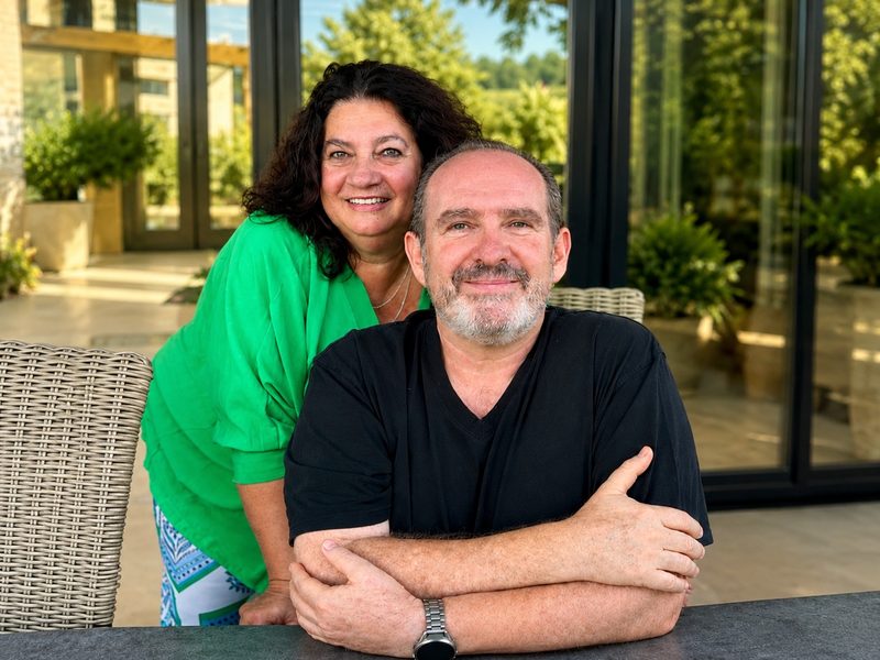 Claus Schertz mit seiner Partnerin, beide lächeln in die Kamera, im Hintergrund eine sonnige Terrasse